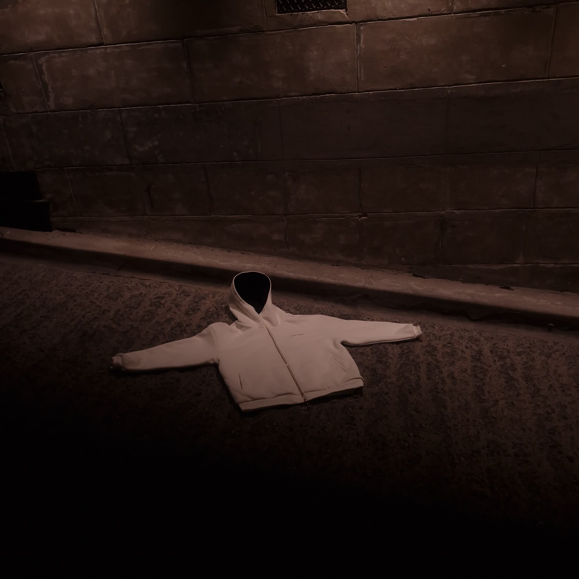Cream hoodie laid flat against a stone-block wall under a single overhead light
